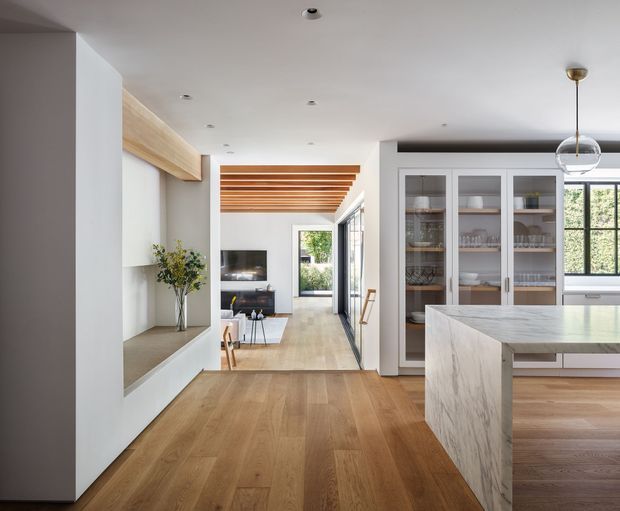 Bright open-plan residential interior showing a contemporary kitchen and living area. The foreground features a large pale stone island with waterfall sides, warm timber flooring, white walls, and built-in glass-fronted display cabinetry. A timber slatted ceiling feature leads toward a compact living area with glazing and garden views. A spherical pendant light and recessed ceiling lights complete the refined, warm space.