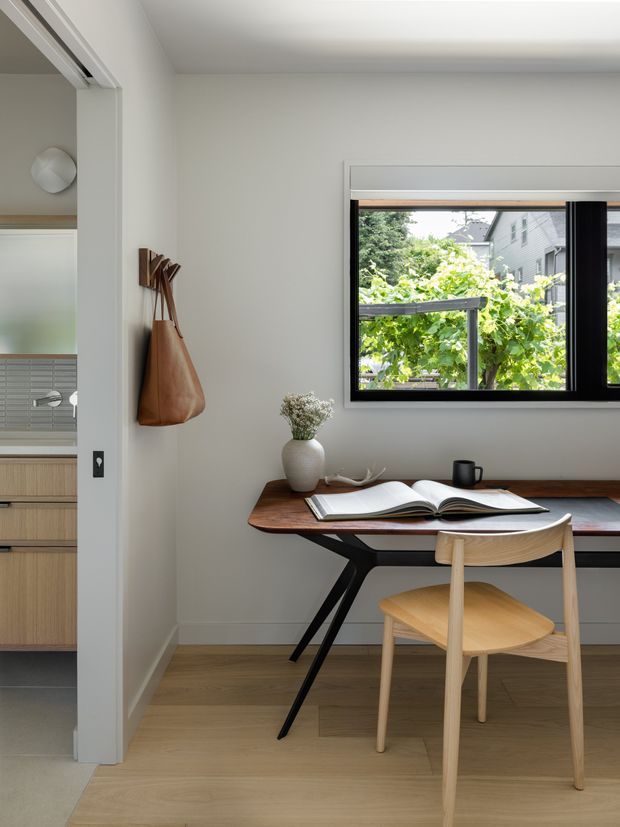 A compact, serene home office with a timber desk, light wooden chair, open book, ceramic vase, and black mug. The room has pale walls, warm timber flooring, a large black-framed window overlooking leafy greenery, and a partial view of a minimalist kitchen through the adjacent opening. A leather bag hangs on wall hooks, adding a small functional accent.