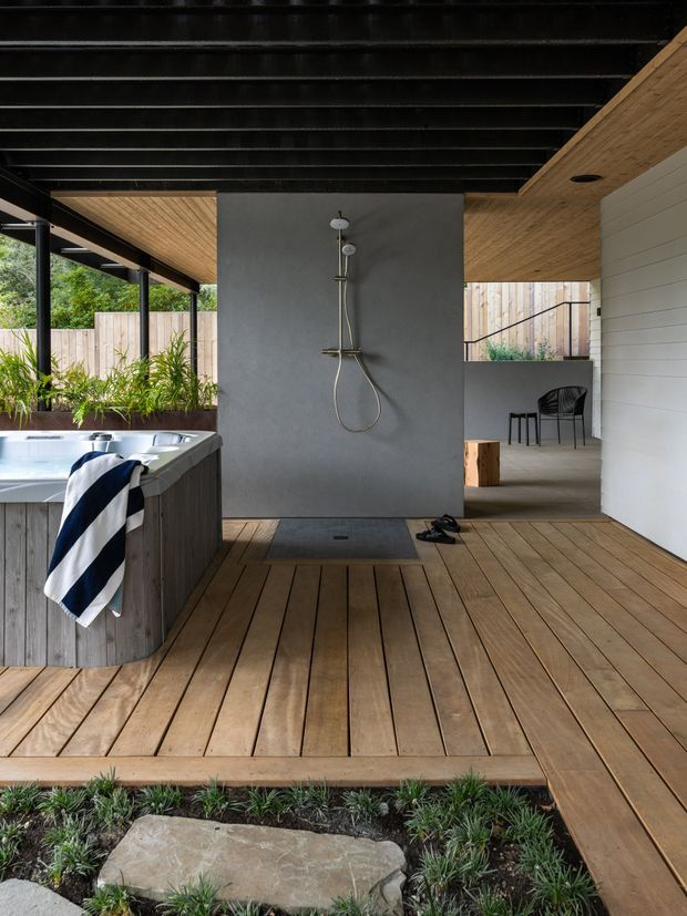 A contemporary outdoor bathing and relaxation area with a timber deck, freestanding spa, and open-air shower mounted on a dark grey tiled or rendered feature wall. Black exposed ceiling beams contrast with warm timber soffits and decking, while a white horizontal-clad wall, timber privacy fencing, integrated planting, and a partially screened seating area frame the space. A stepping stone garden borders the deck.