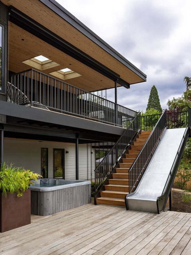 Exterior view of a contemporary multi-level residential home with a broad timber deck, black metal balustrades, open-riser-style timber stairs, and an integrated stainless-steel slide alongside the staircase. The upper level has a timber-lined soffit with skylights and extensive outdoor circulation, while the lower level includes a freestanding outdoor spa clad in vertical timber. Lush planting surrounds the deck and the house.