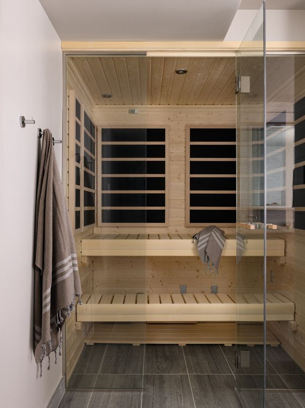 A compact cedar-lined sauna viewed through a frameless glass door. The sauna has two-tier timber benches, horizontal black-backed slatted panels on the walls, a matching timber plank ceiling and walls, and dark large-format tiled flooring outside the sauna enclosure. Towels hang on the adjacent white wall.