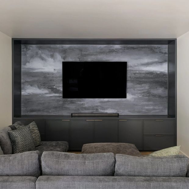 A contemporary media wall viewed from behind a large grey sectional sofa. The wall features a black-framed built-in television niche, a dramatic mottled grey feature surface, and extensive dark built-in cabinetry with drawers and a soundbar. Neutral upholstered seating and cushions occupy the foreground.