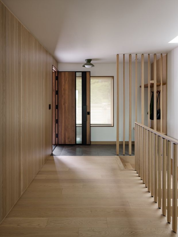 A warm, minimalist residential entry hall or landing with extensive vertical timber wall panelling, light timber flooring, a dark timber front door, and a large window at the far end. Vertical timber slats form a screen and balustrade around the stair opening on the right, with a small coat-hanging area behind them. The space is softly lit by daylight and a simple ceiling light.