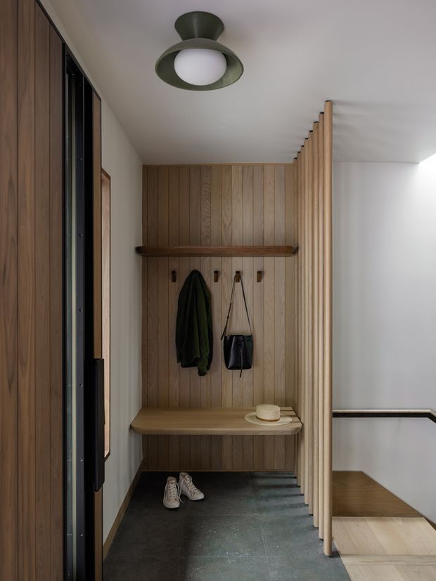 A compact, carefully detailed entryway with full-height vertical timber panelling and matching built-in bench and shelf. Wall hooks hold coats and a handbag, with shoes on a dark tiled floor. A slatted timber screen partially separates the entry from the adjacent stairway, and a sculptural green ceiling light provides a focal point.
