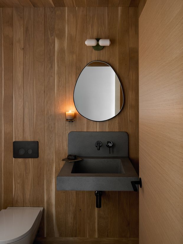 Compact, moody bathroom with warm vertical timber wall panelling, a dark concrete or stone wall-mounted vanity with integrated rectangular basin, black wall-mounted tapware, an organic oval mirror, and warm decorative lighting. A toilet is partially visible at the lower left, while a timber door or partition occupies the right side.
