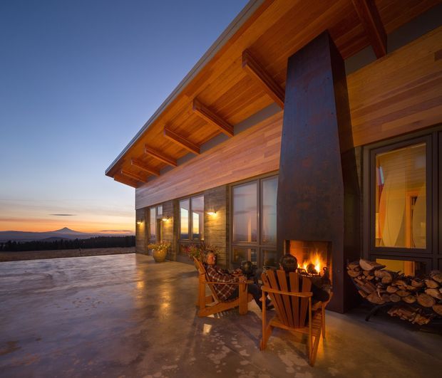 Exterior dusk view of a modern timber-clad residence with a broad covered terrace, exposed timber roof structure, large glazed doors and windows, and a tall dark metal outdoor fireplace with a visible fire. Wooden lounge chairs and stacked firewood occupy the terrace, with a distant mountain landscape beyond.