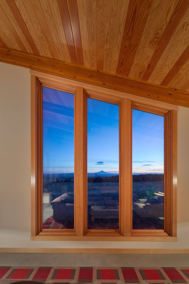Interior view dominated by a tall three-panel timber-framed window overlooking a distant mountain landscape at dusk. The room features warm honey-toned wood trim and a richly patterned exposed timber ceiling with beams. A geometric red, brown, and beige carpet is visible at the bottom of the image, with reflected furnishings seen in the glazing.