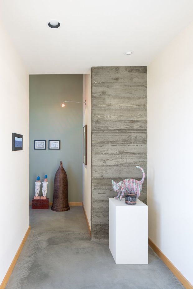 A minimalist interior hallway or entry gallery with polished concrete flooring, white walls, and a prominent vertical textured concrete feature wall. The space displays framed artwork, sculptural objects, and a colourful cat sculpture on a white pedestal. Recessed ceiling lights and a small wall light provide illumination, while a muted sage-green alcove adds contrast.