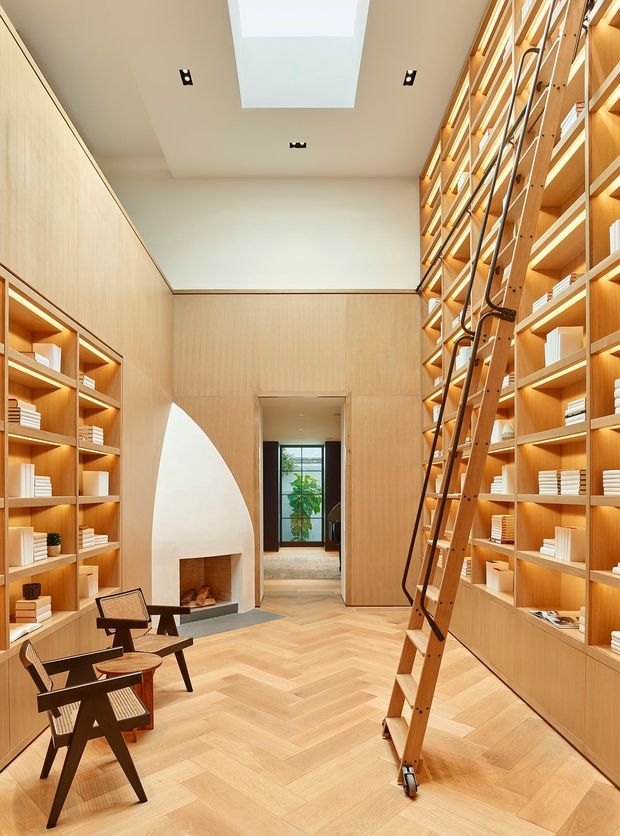 A tall, warm-toned library or reading room lined with floor-to-ceiling light timber shelving and cabinetry, filled with books and reached by a rolling timber ladder. The room has a pale herringbone timber floor, timber wall panelling, a sculptural white fireplace, two dark wood and woven lounge chairs, recessed ceiling lighting, and a large rectangular skylight. A doorway leads to a secondary room with glazing and greenery.