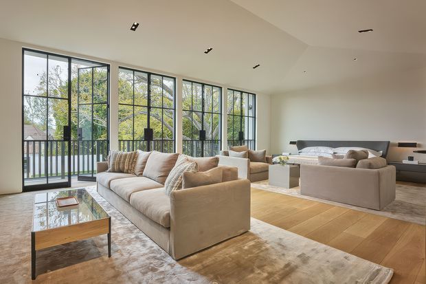 A spacious, bright open-plan living room with a large beige sectional sofa, matching armchairs, a glass-topped coffee table, layered neutral rugs, and warm timber flooring. Full-height black-framed glazing opens to a balcony and provides views of trees. A bedroom area with a bed is visible beyond the seating zone, reinforcing the open-plan residential layout. Recessed ceiling spotlights and a restrained neutral palette create a refined contemporary atmosphere.