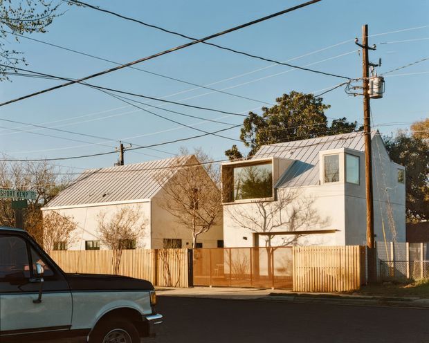 Street view of a compact contemporary residential building with a light metal pitched roof, white rendered walls, projecting box windows, and a timber privacy fence. A neighboring low-rise structure, utility poles and overhead wires, mature trees, and a parked vehicle are visible in the suburban streetscape.