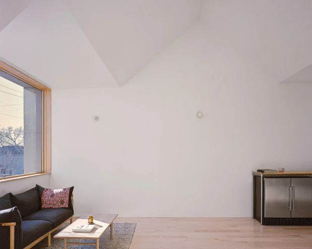Bright, sparsely furnished modern interior with a pale timber floor, white walls and an angular vaulted ceiling. A large timber-framed window brings in natural light beside a dark sofa and patterned cushion, while a low coffee table and compact dark built-in cabinet occupy the room. The space feels calm and minimalist, with an understated contemporary fit-out.