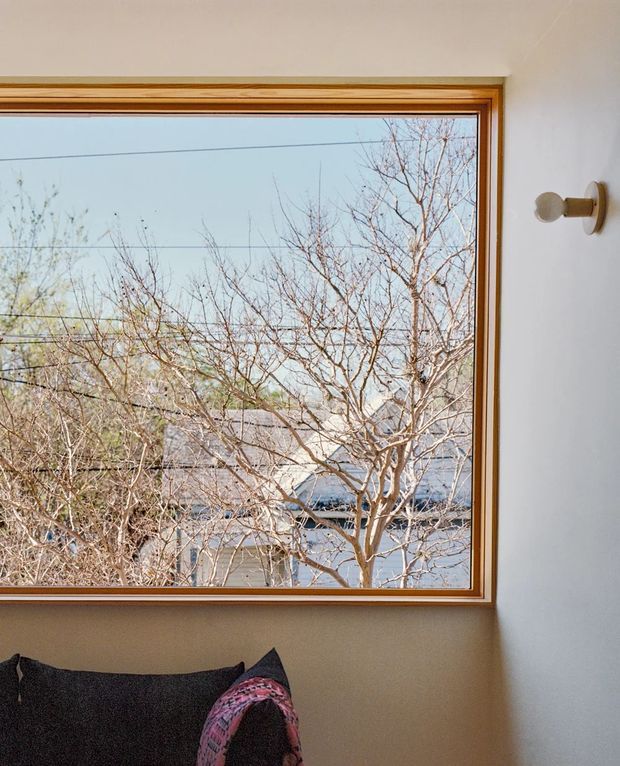 Interior detail centered on a large timber-framed picture window overlooking leafless trees and neighbouring rooftops. The window is set in a plain light wall, with a small wall-mounted cylindrical light fixture to the right and the corner of a dark upholstered sofa with a patterned cushion at the bottom. The scene has a restrained, warm, natural palette and simple contemporary detailing.