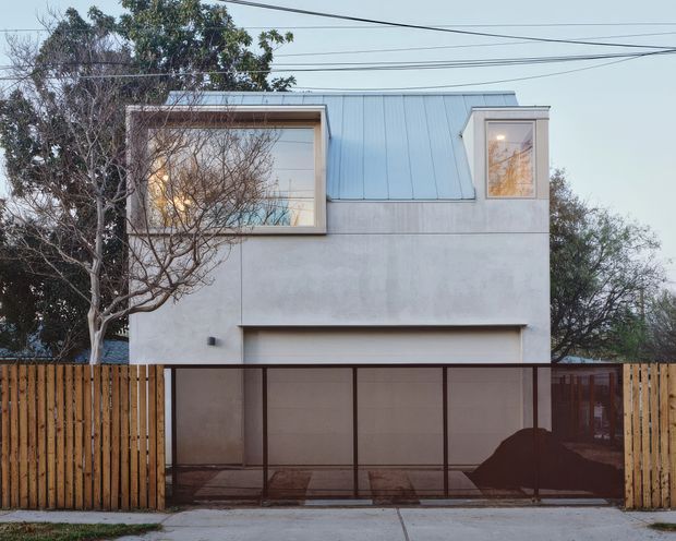 Street-facing view of a compact contemporary two-storey residence. The facade combines smooth pale rendered walls with a standing-seam metal upper roof form and projecting box-framed windows. A wide dark-framed garage or carport opening is screened by a translucent panel gate, while timber paling fencing encloses the property. Mature trees and overhead utility wires surround the building.