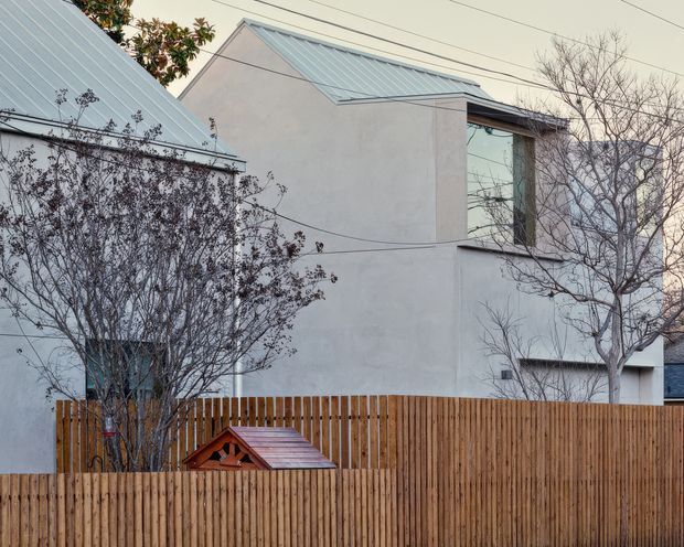 Exterior view of a contemporary multi-level residence with smooth pale rendered walls, simple angular volumes, standing-seam metal gabled roofs, and a tall glazed window. A timber vertical-slat fence and small timber garden structure occupy the foreground, with leafless trees partially screening the building.