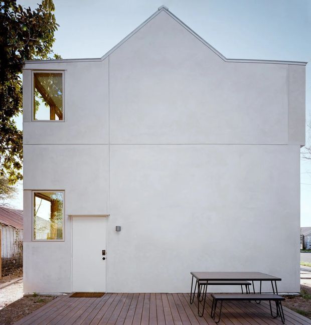 Minimal two-storey white rendered house facade with a simple gabled roof profile, tall rectangular windows, and a plain entry door. A timber deck and outdoor picnic table occupy the foreground, with neighboring structures and trees at the edges.