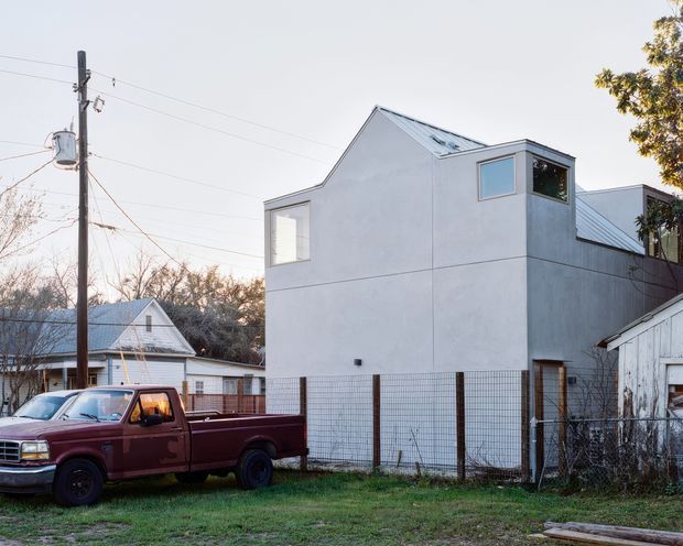 Exterior view of a contemporary, narrow multi-level residential building with a simple white rendered facade, prominent gabled roof forms, and projecting upper-level window volumes. The property is enclosed by a wire-mesh fence and sits in a modest semi-rural or suburban setting, with older neighboring buildings and a parked pickup truck in the foreground.