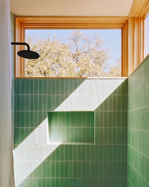 A bright, compact shower room with deep green vertically arranged wall tiles and a contrasting white tiled geometric niche. A black wall-mounted rain shower is visible on the left. The shower is framed by warm timber window and wall trim, with a large open window overlooking a leafy tree and allowing strong sunlight to cast angular shadows across the tiled surfaces.