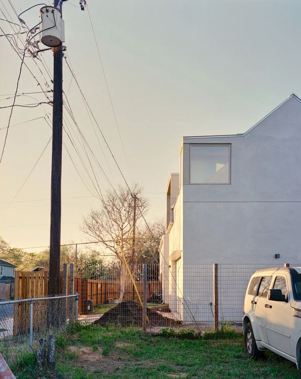 Exterior view of a contemporary multi-level residential building beside a fenced yard and driveway. The building has smooth pale rendered walls, simple rectangular glazing, and an asymmetrical pitched/gabled roofline. Utility poles and overhead wires dominate the streetscape, with a parked van visible in the foreground.