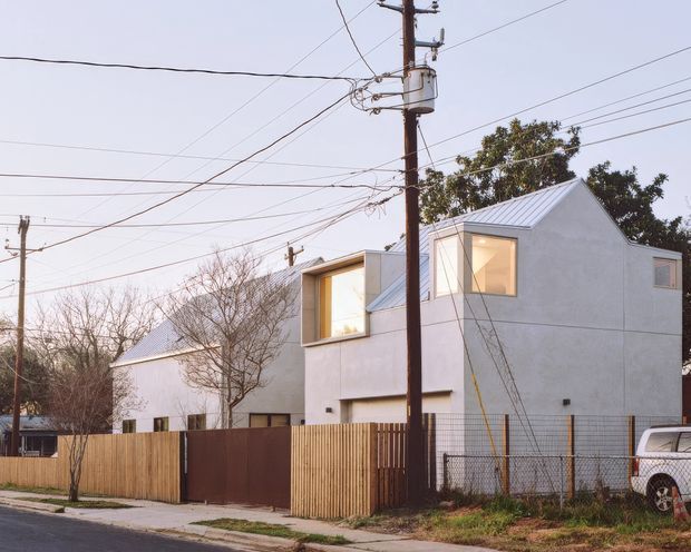 Street-level exterior view of a contemporary multi-storey residential building with crisp pale rendered walls, steep gabled metal roofs, projecting box-like upper-level windows, and a timber privacy fence. Utility poles and overhead wires dominate the foreground, with neighbouring structures, bare trees, and a parked vehicle visible around the property.
