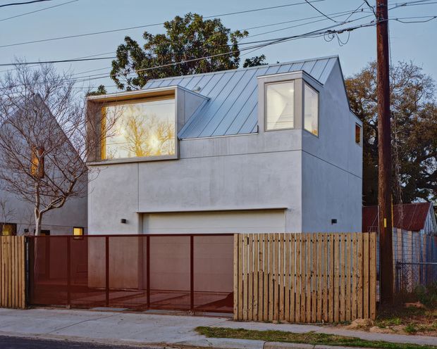 Street-facing view of a compact contemporary two-storey house with a minimalist pale rendered facade, angular standing-seam metal roof forms, large projecting glazed window, and upper-level corner windows. A translucent red metal vehicle gate and vertical timber fence define the frontage, with overhead utility wires and mature trees surrounding the property.