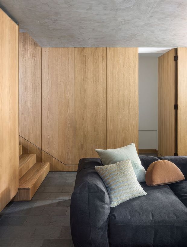 A refined contemporary interior showing a dark charcoal sectional sofa in the foreground, surrounded by extensive vertical light-oak wall panelling and concealed cabinetry. A compact timber stair rises along the left wall, while the floor is finished in dark grey stone or slate tiles. The ceiling has a raw, textured concrete finish, creating a warm yet restrained material palette.