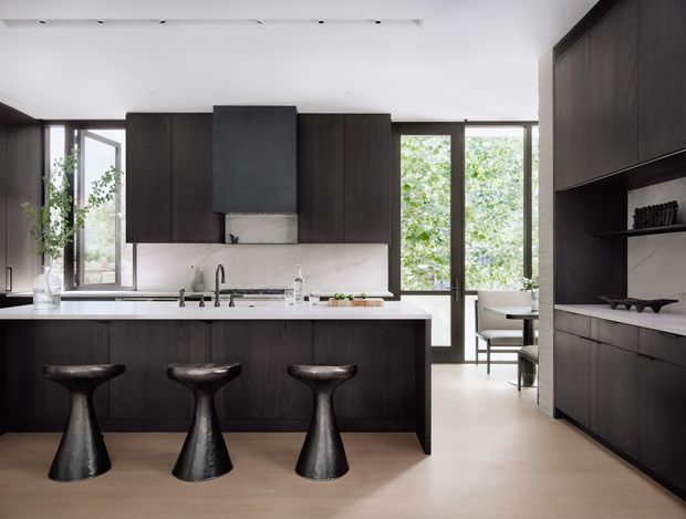 Contemporary kitchen with a large dark timber island, three sculptural black bar stools, white stone benchtops, dark full-height cabinetry, and a pale stone backsplash. Full-height glazed doors and windows bring in leafy views and natural light, while a dining area is visible beyond the kitchen. Integrated linear ceiling lighting and minimalist detailing create a refined, high-contrast interior.