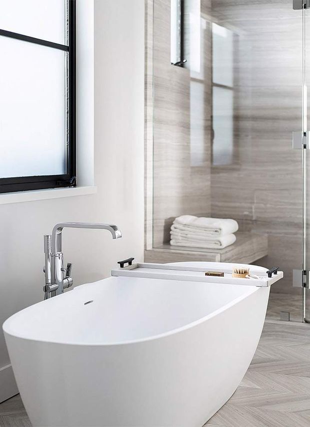 Bright contemporary bathroom featuring a freestanding white bathtub, chrome floor-mounted tapware, a frameless glass shower enclosure with a built-in tiled bench, and folded towels. The room has pale stone-look wall tiles, light herringbone-pattern floor tiles, white walls, and black-framed window detailing.