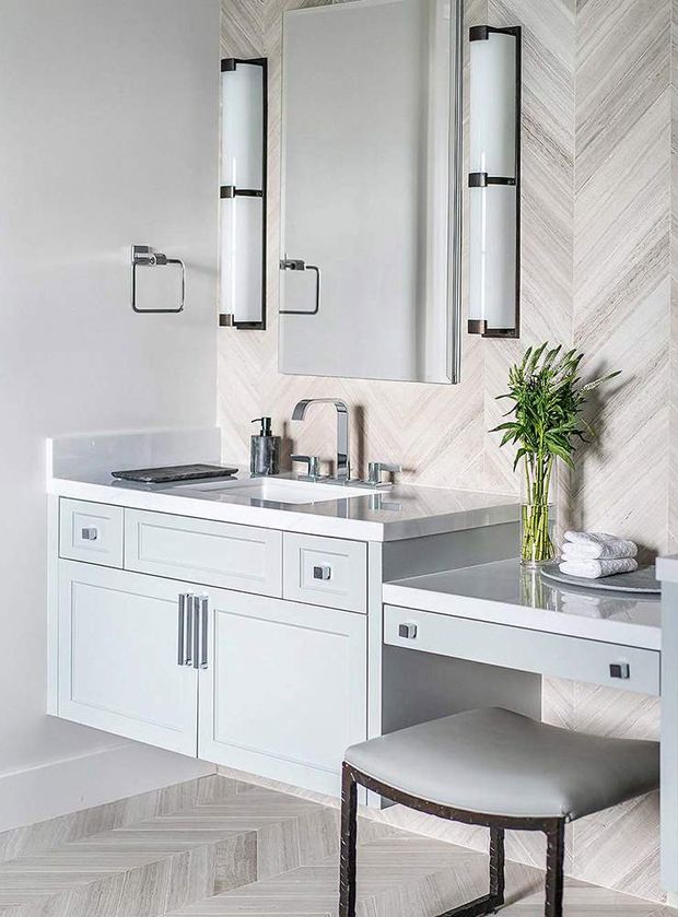 Bright, refined bathroom vanity area with white built-in cabinetry, a broad rectangular mirror, twin vertical wall sconces, a polished light-grey countertop with integrated double basins, and brushed metal fixtures. The walls and floor feature pale herringbone-patterned tile, while a floating side vanity or makeup desk extends to the right with a cushioned stool. Fresh greenery and folded towels add softness.