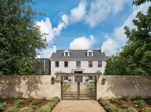 A symmetrical, traditional two-storey residence viewed from the front through a gated landscaped approach. The pale rendered facade has a dark grey pitched roof with dormer windows, white chimneys, black-framed multi-pane windows, and a small classical balcony. A tan brick boundary wall, metal driveway gate, paved path, and formal garden beds frame the entrance.