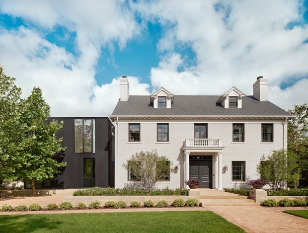 Front exterior view of a large two-storey brick residence with a symmetrical traditional facade, dark pitched roof, dormer windows, chimneys, and a columned entrance porch. A contrasting dark contemporary wing extends from the left side. The landscaped frontage includes a lawn, paved path, low hedging, and mature trees under a partly cloudy sky.