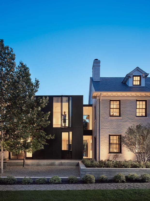 Twilight exterior view of a residential property combining a restored traditional brick house with a contrasting black, box-like contemporary extension. The original house has a pitched slate roof, prominent chimney, dormer window, decorative eaves, and multi-pane windows, while the extension features dark cladding and tall glazed openings. A landscaped front garden and gravel forecourt sit in front of the building.
