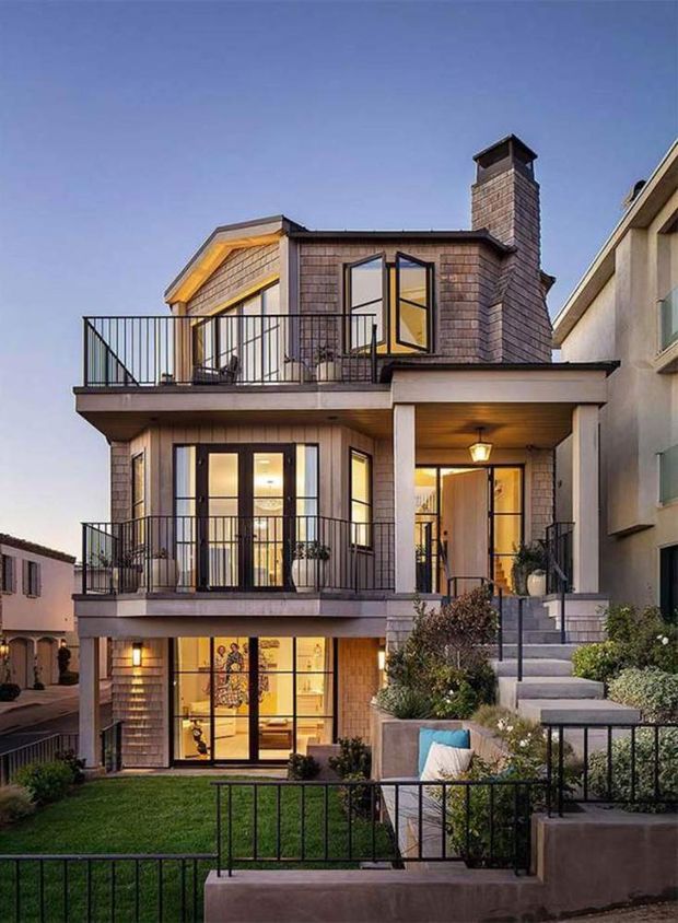 Exterior view of a narrow multi-storey urban residence at dusk, featuring shingle-clad upper walls, multiple balconies with slim black metal railings, large glazed doors and windows, a prominent masonry chimney, and a covered entry porch reached by steps. Landscaped terraces, lawn, built-in seating, and neighbouring homes surround the facade.