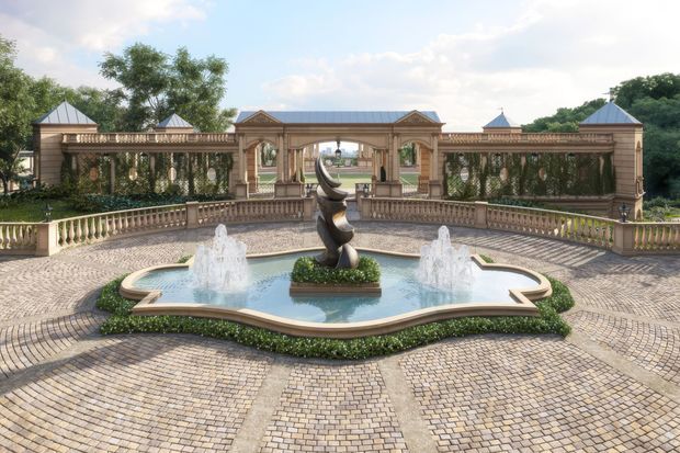 Formal landscaped courtyard with a central sculptural fountain pool, symmetrical water jets, ornate stone balustrades, and a classical colonnaded garden pavilion with multiple small pitched roof elements. The ground is paved in patterned light cobblestones, with clipped hedges and mature trees surrounding the grand garden composition.