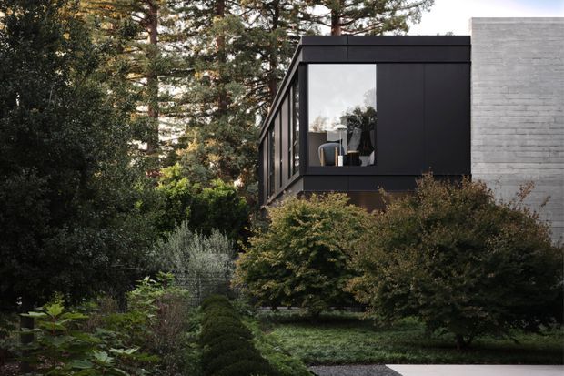 Exterior view of a contemporary dark-clad building extension or wing set within a lush, mature landscaped garden. The structure features black rectangular cladding, large floor-to-ceiling windows, and a contrasting pale grey-white brick wall. Dense trees, shrubs, sculpted greenery, and a paved path frame the building.