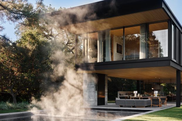 Contemporary two-storey house set within a landscaped garden, featuring a dark rectilinear upper volume, extensive floor-to-ceiling glazing, warm timber soffits, and a partially open ground-level living and dining area. Steam rises from a linear pool or water feature in the foreground, adding a dramatic atmospheric element.