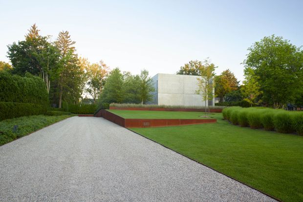 A landscaped contemporary residential exterior viewed from a broad pale gravel driveway. A minimalist rectangular concrete building volume sits beyond a geometric raised garden bed edged in rust-red metal or masonry, surrounded by manicured lawn, clipped hedges, ornamental grasses, and mature trees.