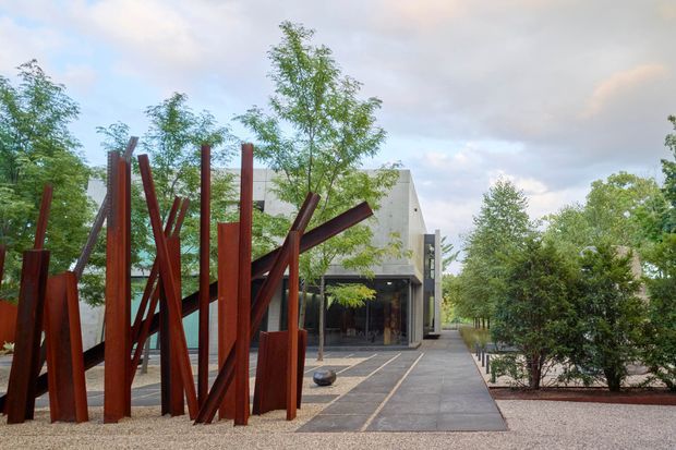 A contemporary building entrance viewed through a landscaped forecourt, with a large sculptural installation of rust-coloured steel beams in the foreground. The building has a geometric light-coloured facade, dark glazed entry, paved walkways, gravel landscaping, and mature trees surrounding the approach.