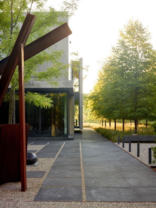 A modern architectural exterior viewed from a landscaped garden, featuring a minimalist concrete-and-glass building, dark stone paving, gravel beds, slender bollards, mature trees, and a prominent rusted steel sculptural structure in the foreground. The composition has strong geometric lines and a tranquil, natural setting.