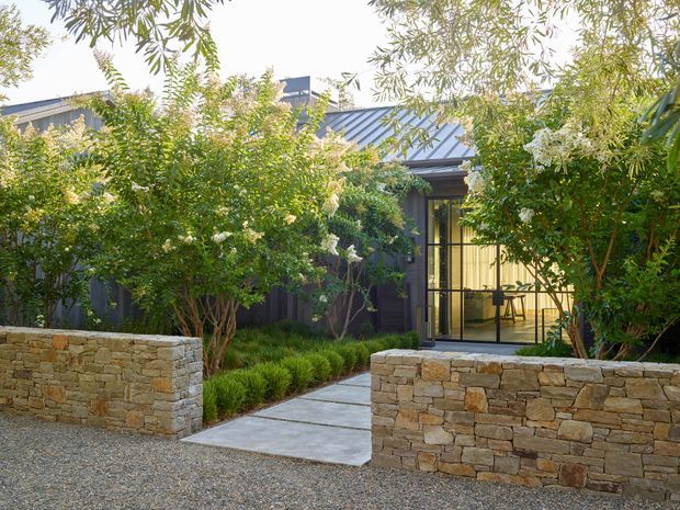 A landscaped courtyard entry to a dark-clad, low-profile building. The approach is framed by rough stacked-stone retaining walls, gravel paving, concrete stepping slabs, dense ornamental planting, and a glazed black-framed entrance beneath a metal pitched roof. Warm interior light and a small table are visible through the glass.