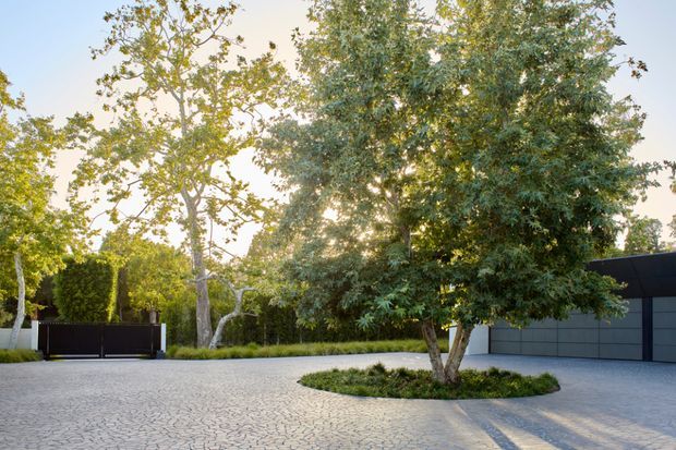 A landscaped residential driveway or forecourt with pale irregular-pattern paving, mature trees, clipped hedges, and a dark contemporary boundary gate and garage facade. The circular tree island and broad paved approach create a calm, carefully designed exterior arrival space.