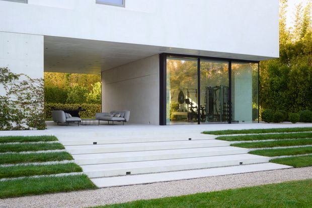 Modern minimalist residential exterior with a white rectangular cantilevered upper volume, expansive dark-framed floor-to-ceiling glazing, and a recessed covered outdoor lounge beneath. Broad pale stone or concrete steps lead through neatly striped lawn and gravel landscaping, while the glazing reveals a home gym inside. The composition is clean-lined, bright, and highly geometric.