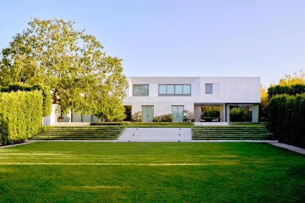 Front exterior view of a modern two-storey white residence set behind a broad manicured lawn. The house has a minimalist rectangular composition, flat roof, smooth pale facade, large horizontal windows, deep glazed openings, and broad tiered steps leading up from the lawn. Tall clipped hedges frame the property, with a mature tree on the left.