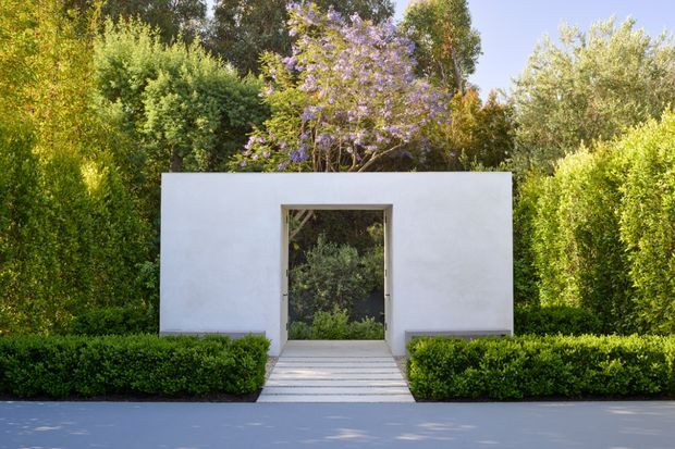A minimalist white garden structure or pavilion set within a formally landscaped garden. The rectangular rendered facade has a single large glazed doorway or opening framed in a darker material, reached by broad pale steps. Dense clipped hedges and mature green planting surround the structure, with a purple-flowering tree visible behind it. The image is a frontal exterior architectural photograph in bright natural light.