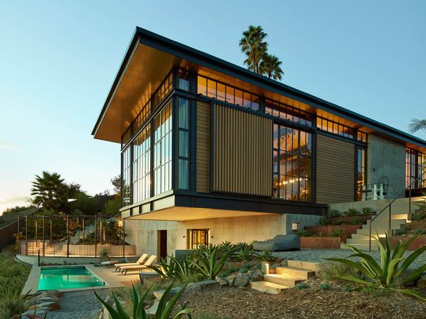 A modern multi-level residence set into a landscaped sloping site, with a dramatic low-pitched roof, extensive floor-to-ceiling glazing, dark metal framing, vertical timber screening, and warm illuminated interiors. Terraced steps lead through drought-tolerant planting and stone landscaping toward a small swimming pool and outdoor lounge area.