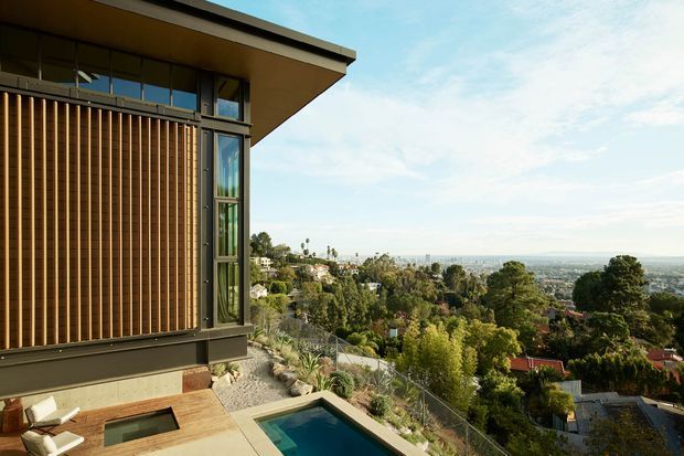 Exterior architectural view of a modern home on a sloping site, showing a projecting flat roof, dark-framed glazing, and a vertical timber screening facade. A terrace with a narrow swimming pool overlooks a densely landscaped suburban valley and distant urban landscape.