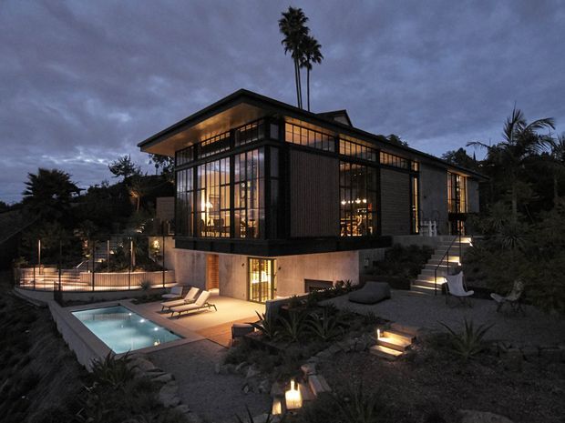 A dramatic dusk exterior view of a contemporary multi-level residence set into a landscaped site. The house features extensive floor-to-ceiling glazing framed in dark metal, a low-pitched roof, dark vertical facade elements, warm interior lighting, terraces and stairways, and a small illuminated swimming pool in the foreground.