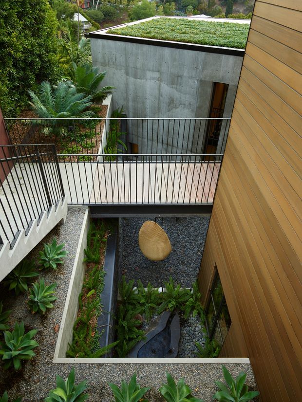 A high-angle view into a landscaped architectural courtyard between contemporary building volumes. The space features raw concrete walls, warm horizontal timber cladding, black metal railings, gravel and pebble ground surfaces, dense planting, and a small dark water feature with a sculptural woven form. A planted green roof is visible on the concrete volume, creating an integrated garden-like setting.