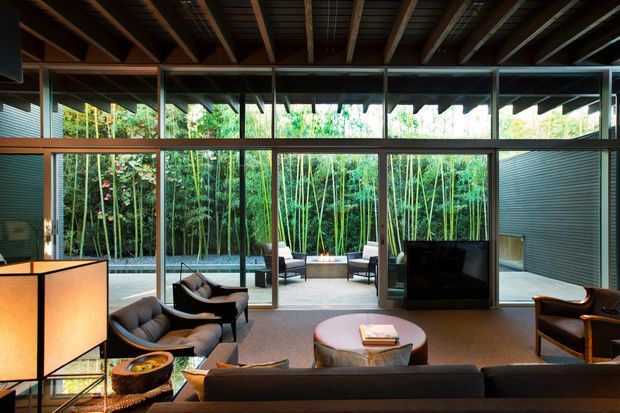 A spacious contemporary living room with dark timber ceiling rafters, extensive floor-to-ceiling glazing, and an open connection to a planted outdoor courtyard. The room contains a large sectional sofa, lounge chairs, a low rounded coffee table, a television, and warm sculptural lighting. Dense bamboo planting is visible beyond the glazing.