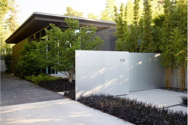 A contemporary single-storey house exterior viewed from the front, with dark horizontal cladding, a broad low-pitched roof, extensive glazing partly screened by planting, and a minimalist white rendered boundary wall and gate. A paved driveway and path lead through dense bamboo and landscaped garden beds.